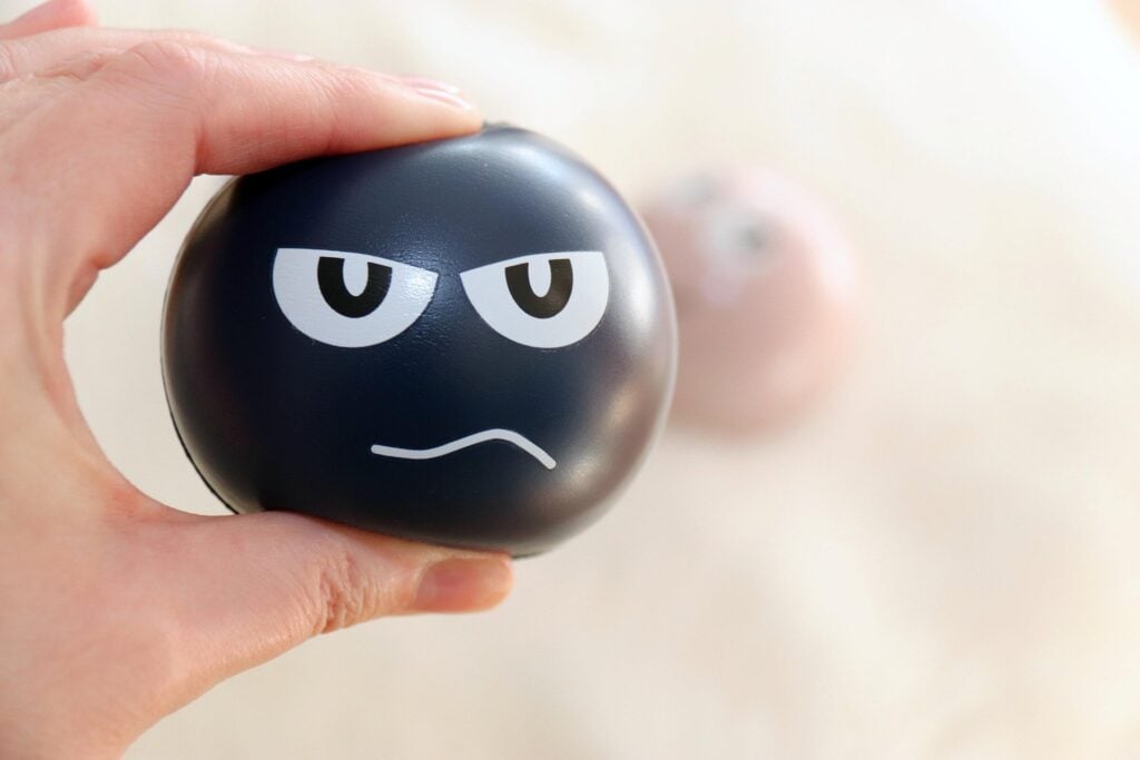A hand squeezing a stress ball, which has a stressed out face on the side of it.