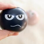 A hand squeezing a stress ball, which has a stressed out face on the side of it.