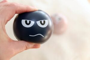 A hand squeezing a stress ball, which has a stressed out face on the side of it.