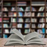 An open book on a table with a large bookshelf in the background slightly blurred.
