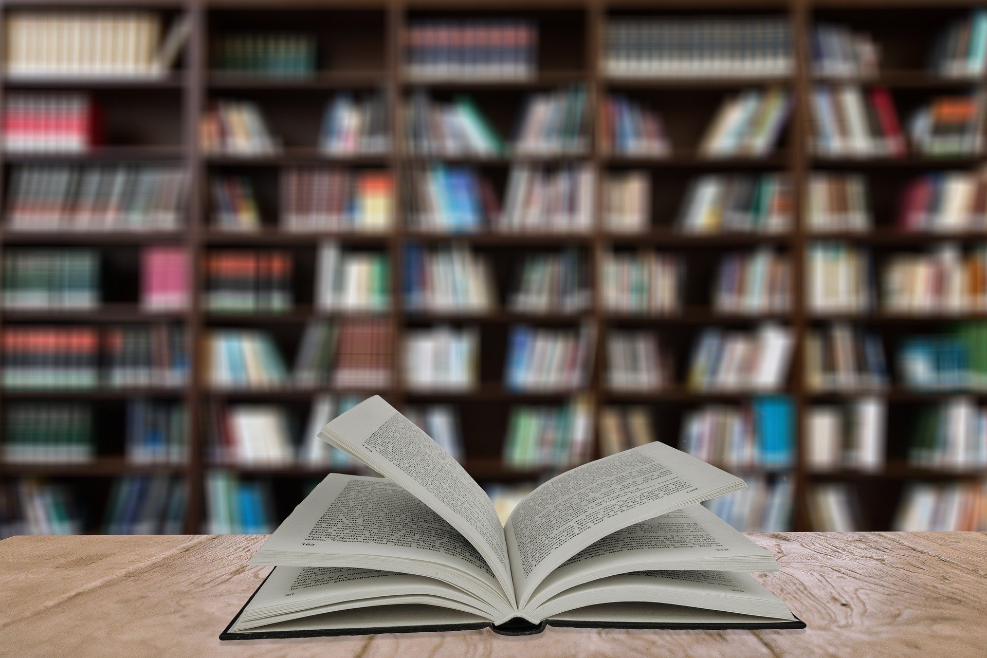 An open book on a table with a large bookshelf in the background slightly blurred.