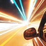A car heading fast down the street, from the driver perspective, with light beams streaming outward towards the highway.