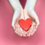 Open hands holding a wooden red heart.