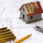 Blueprints with small model of a house on top of it, along with rulers, pencil, pliers.
