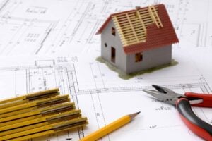 Blueprints with small model of a house on top of it, along with rulers, pencil, pliers.