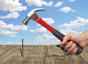 Hammering a nail into a wood deck, blue sky in the background.